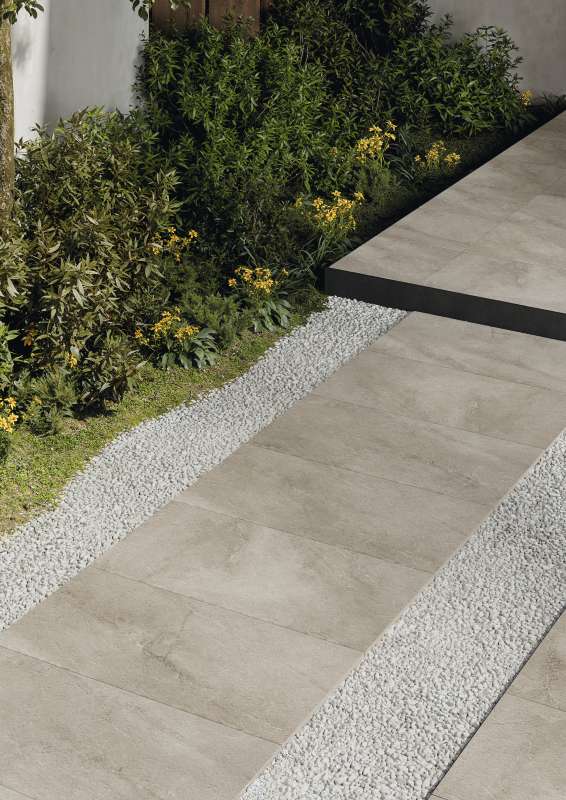 Moderne Outdoor-Fliesen in Grau als Weg und erhöhte Fläche, umgeben von Kies und Grünpflanzen.
